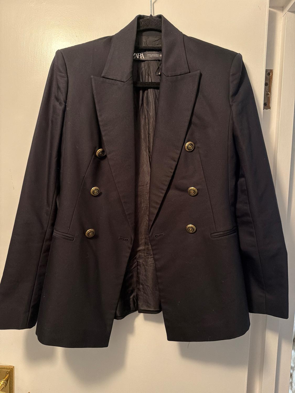 Black double-breasted jacket with gold buttons, hanging on a hanger over a white door.