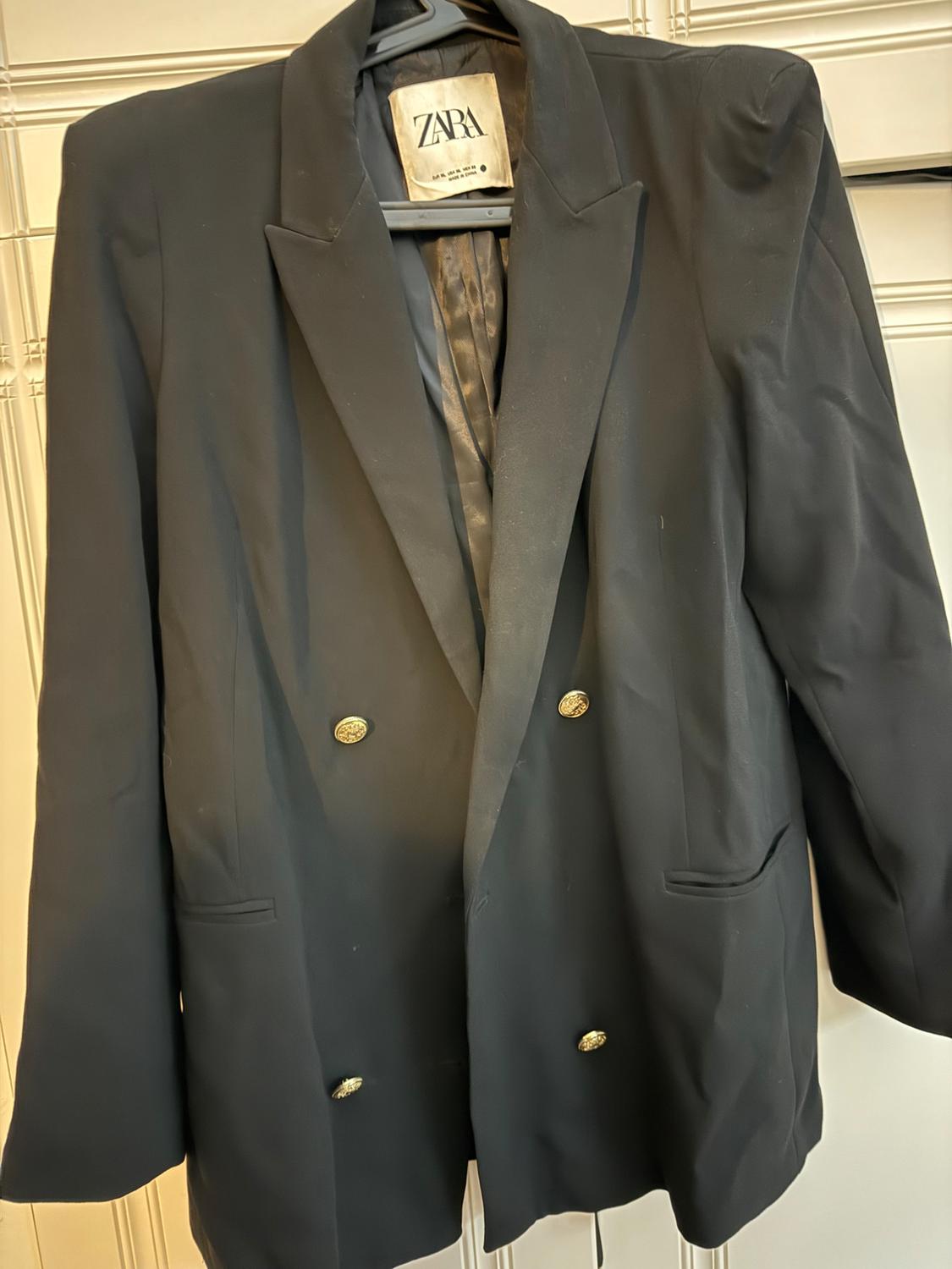 Black double-breasted jacket with gold buttons hanging on a hanger. Visible label with the text "ZARA".