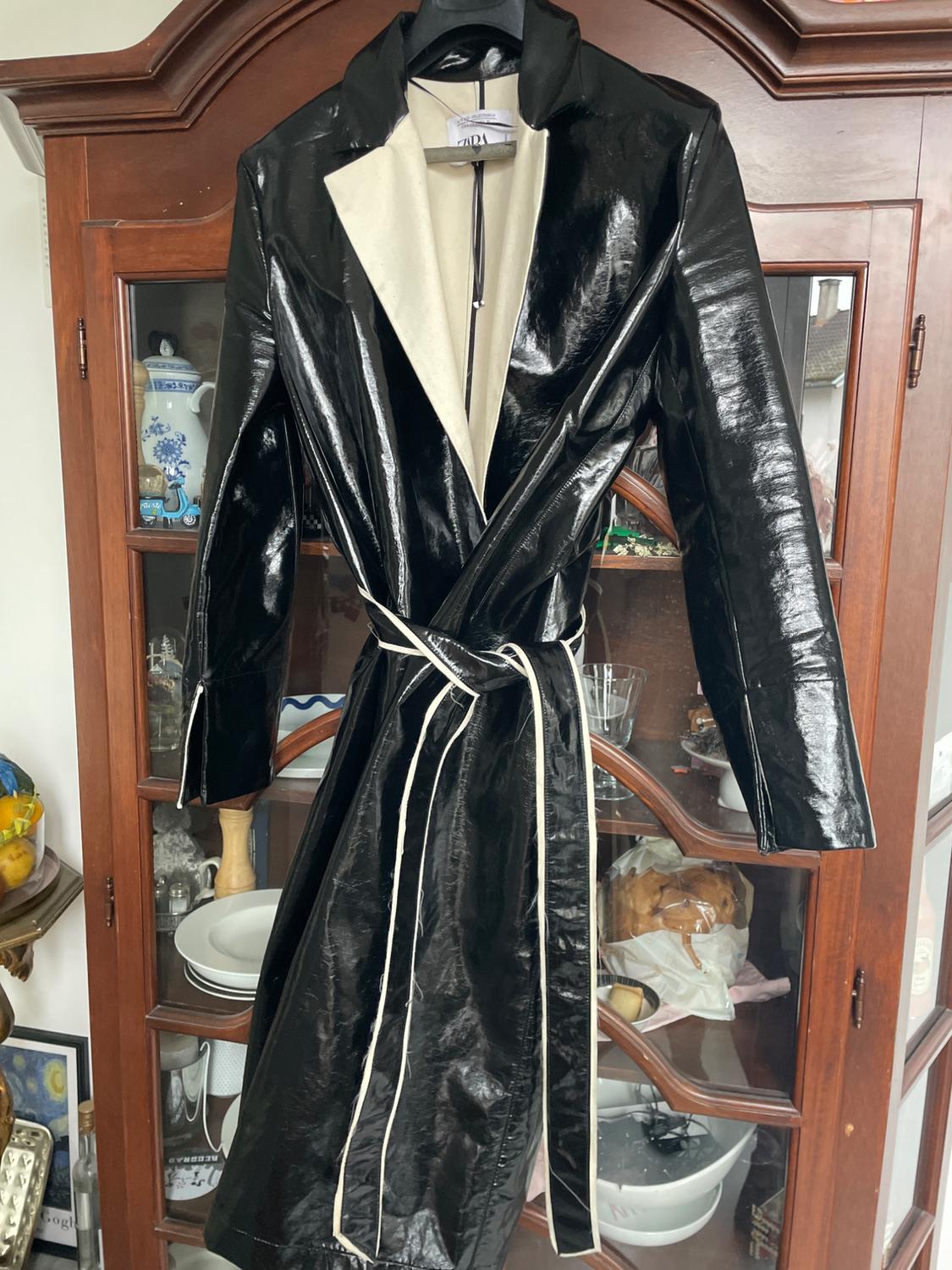 Shiny black coat with a belt and beige lapel hanging in a wooden wardrobe with crockery and decorations in the background.