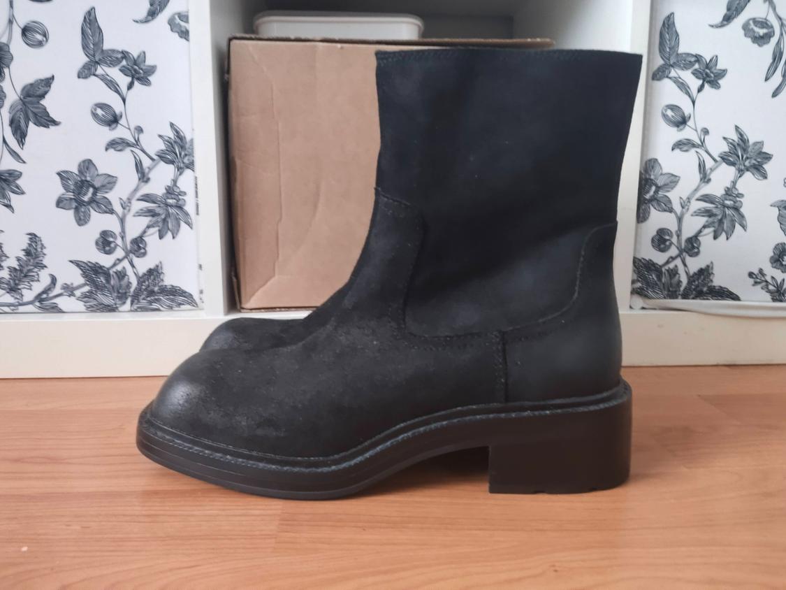 Black low-heeled boot on a wooden floor, with a black and white floral wallpaper background and cardboard box.