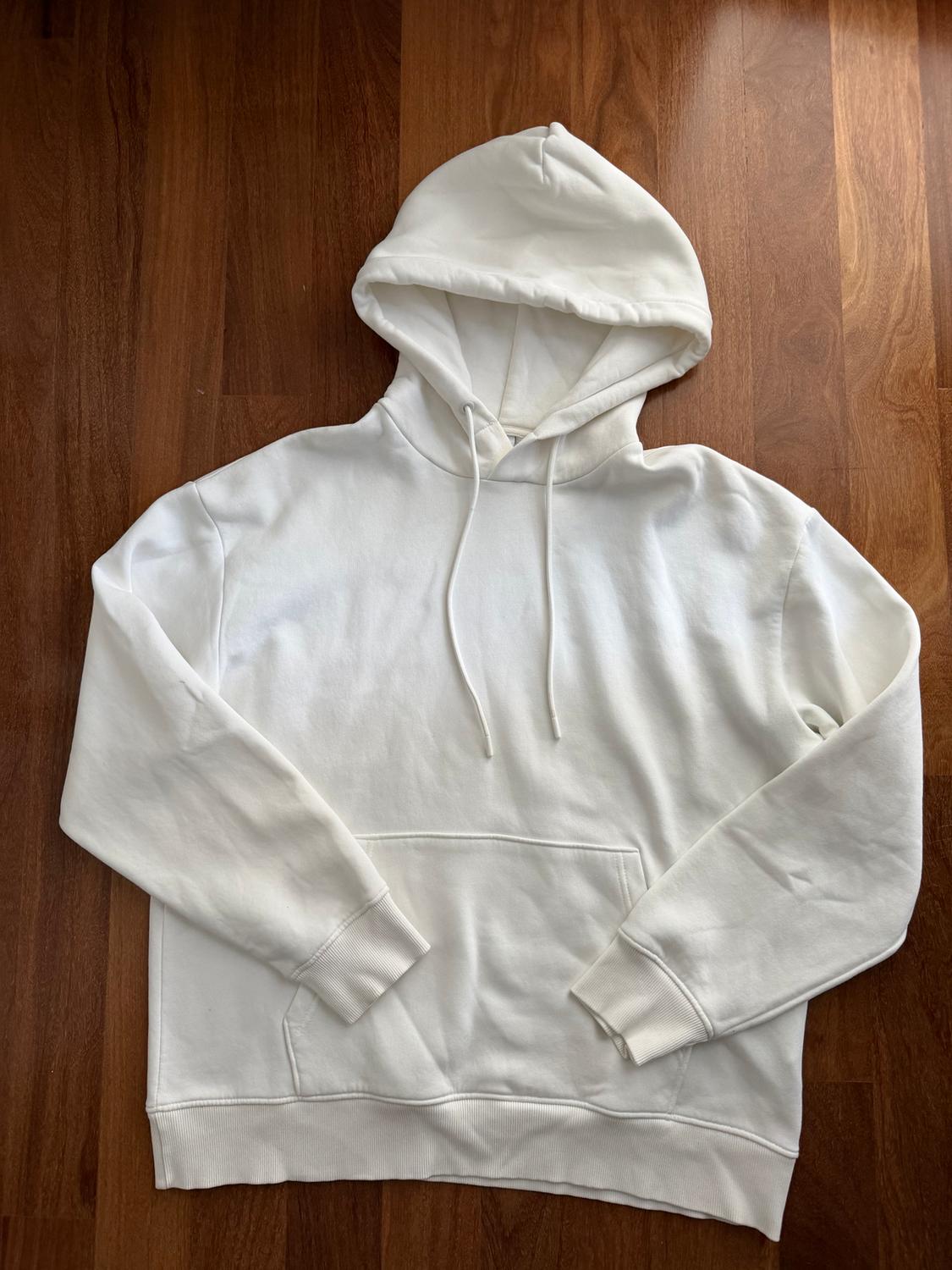 White hoodie with a front pocket, laid out on a brown wooden floor.