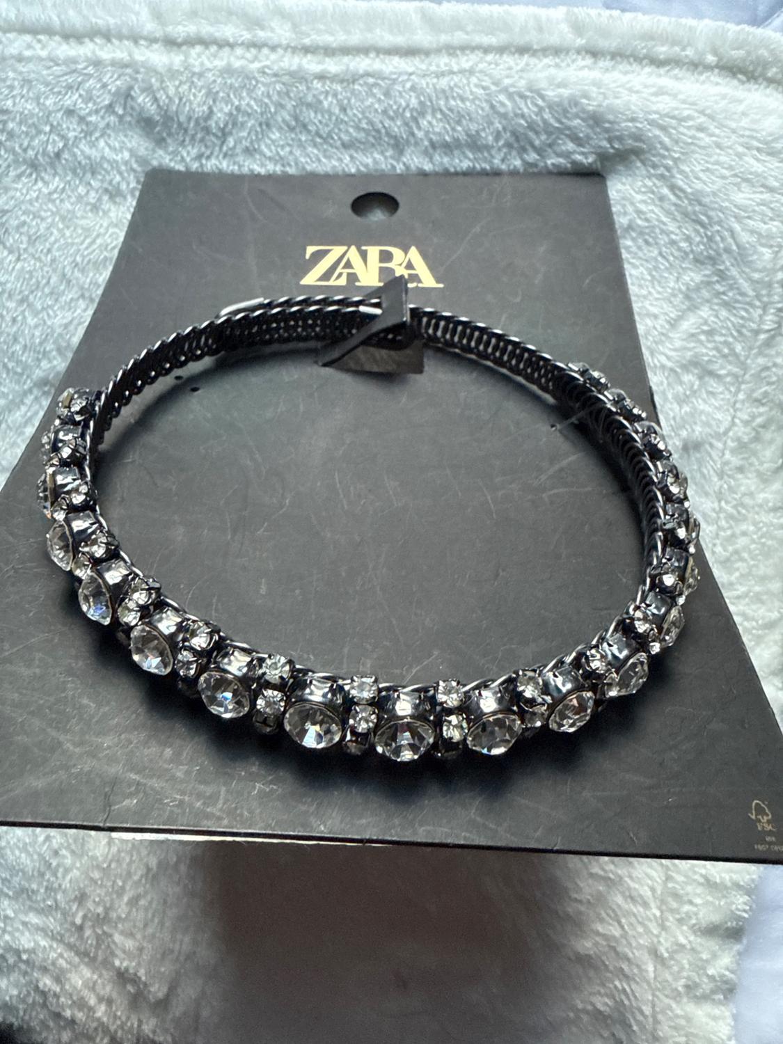 Black headband with rhinestones on a black cardboard background with the "ZARA" logo in gold letters. Light gray fabric background.