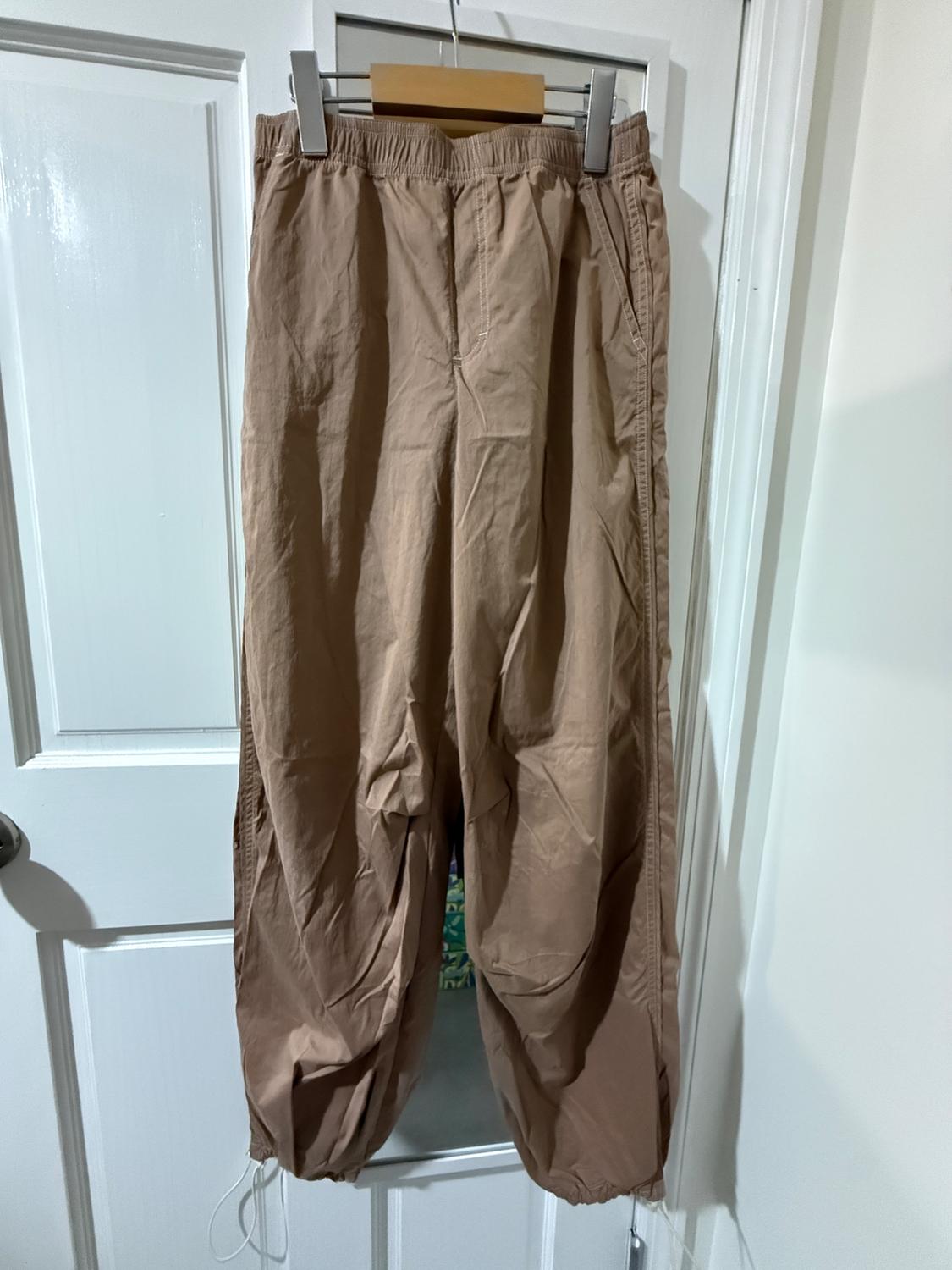Brown trousers hanging on a white door, with an elasticated waist and drawstrings at the ankles. Well-lit indoor setting.