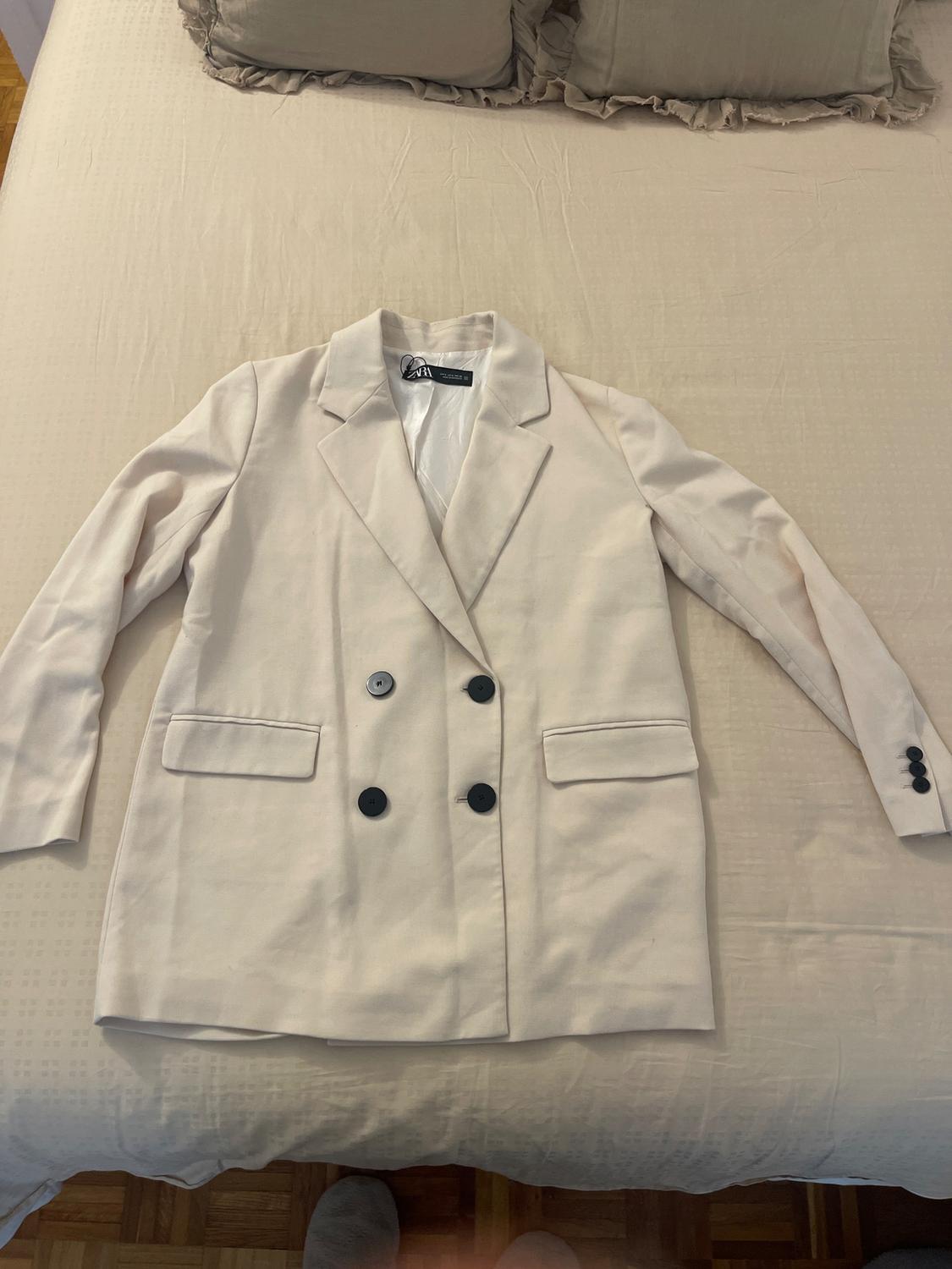 Double-breasted white jacket with black buttons, placed on a bed with a beige bedspread and matching cushions.