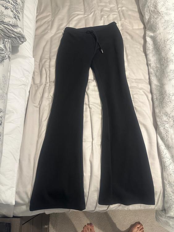 Black flared trousers with a drawstring waist, placed on a bed with white and patterned sheets.