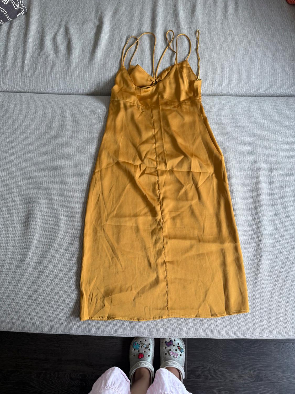 Yellow dress with thin straps on a grey sofa. At the bottom, feet wearing silver clogs and pink trousers are visible.