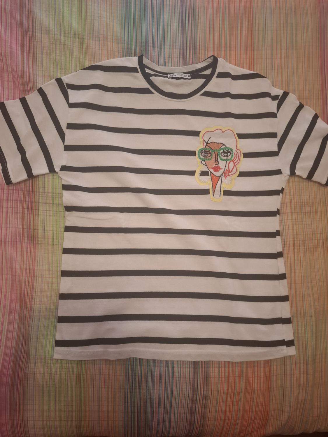 Black and white striped T-shirt with a ZARA label. On the chest, a patch featuring a female face with green glasses. Background of coloured stripes.