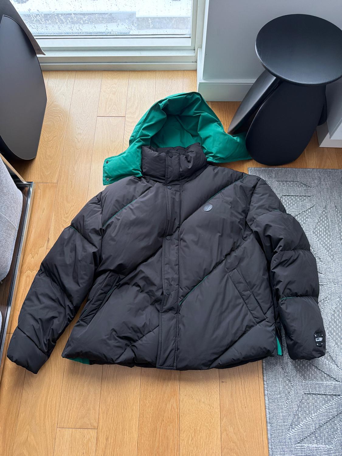 Black padded jacket with a green hood, placed on a wooden floor next to a window and a black table.