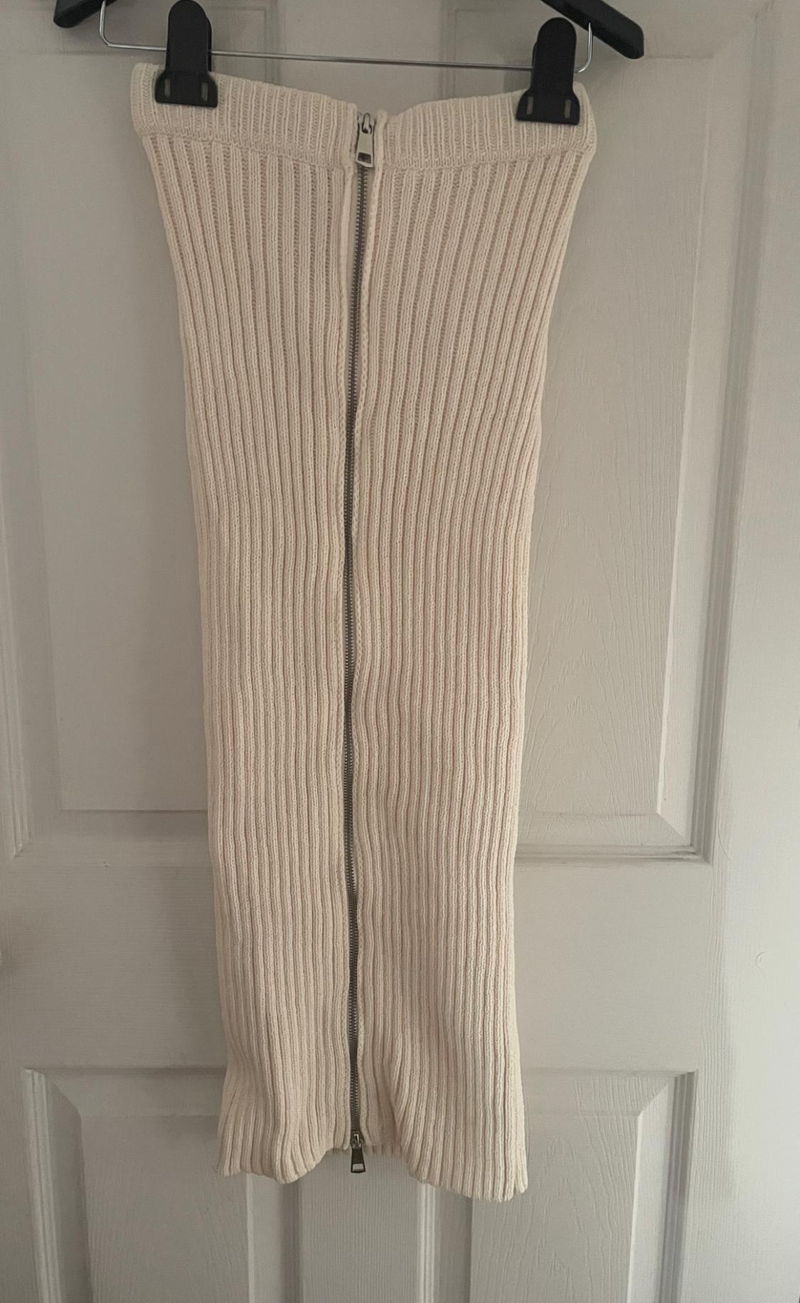 Beige knitted skirt with a central zip, hanging on a black hanger over a white door.