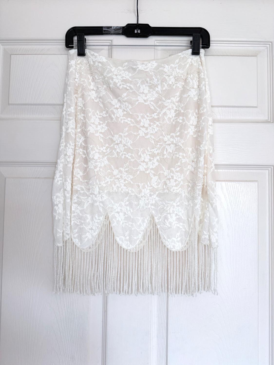 White lace skirt with a scalloped hem and long fringe, hanging from a black hanger on a white door.