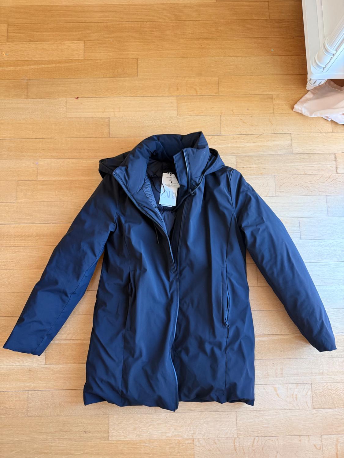 Σκούρο μπλε παλτό με κουκούλα πάνω σε ξύλινο δάπεδο. Διαθέτει μπροστινό φερμουάρ και ετικέτα με το κείμενο ZARA.