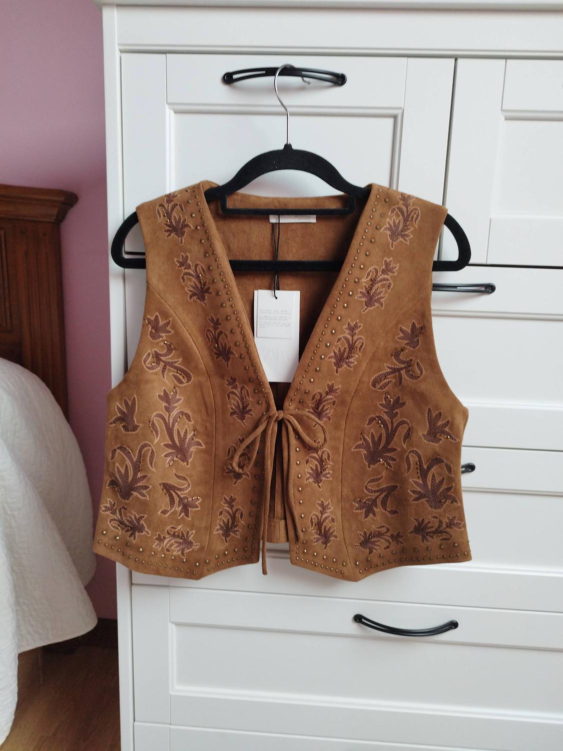 Brown waistcoat with embroidery and studs hanging on a black hanger over a white cabinet in a room with a pink wall.