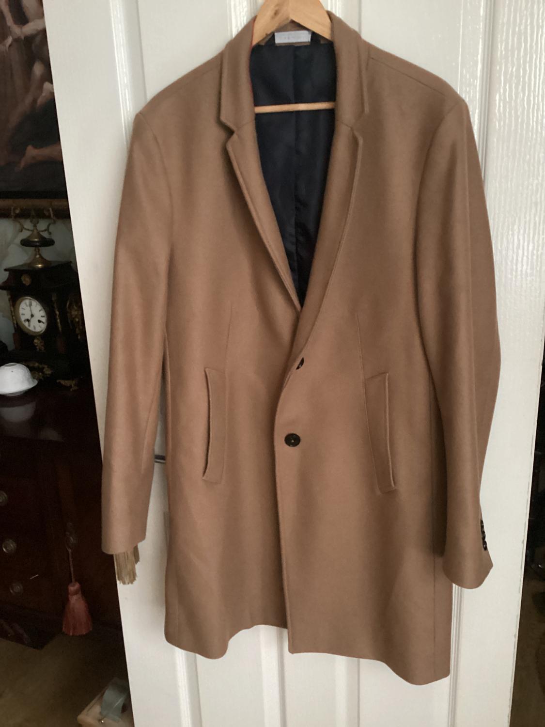 Camel coat hanging on a white door, with a black button and side pockets. Background with dark furniture and an antique clock.