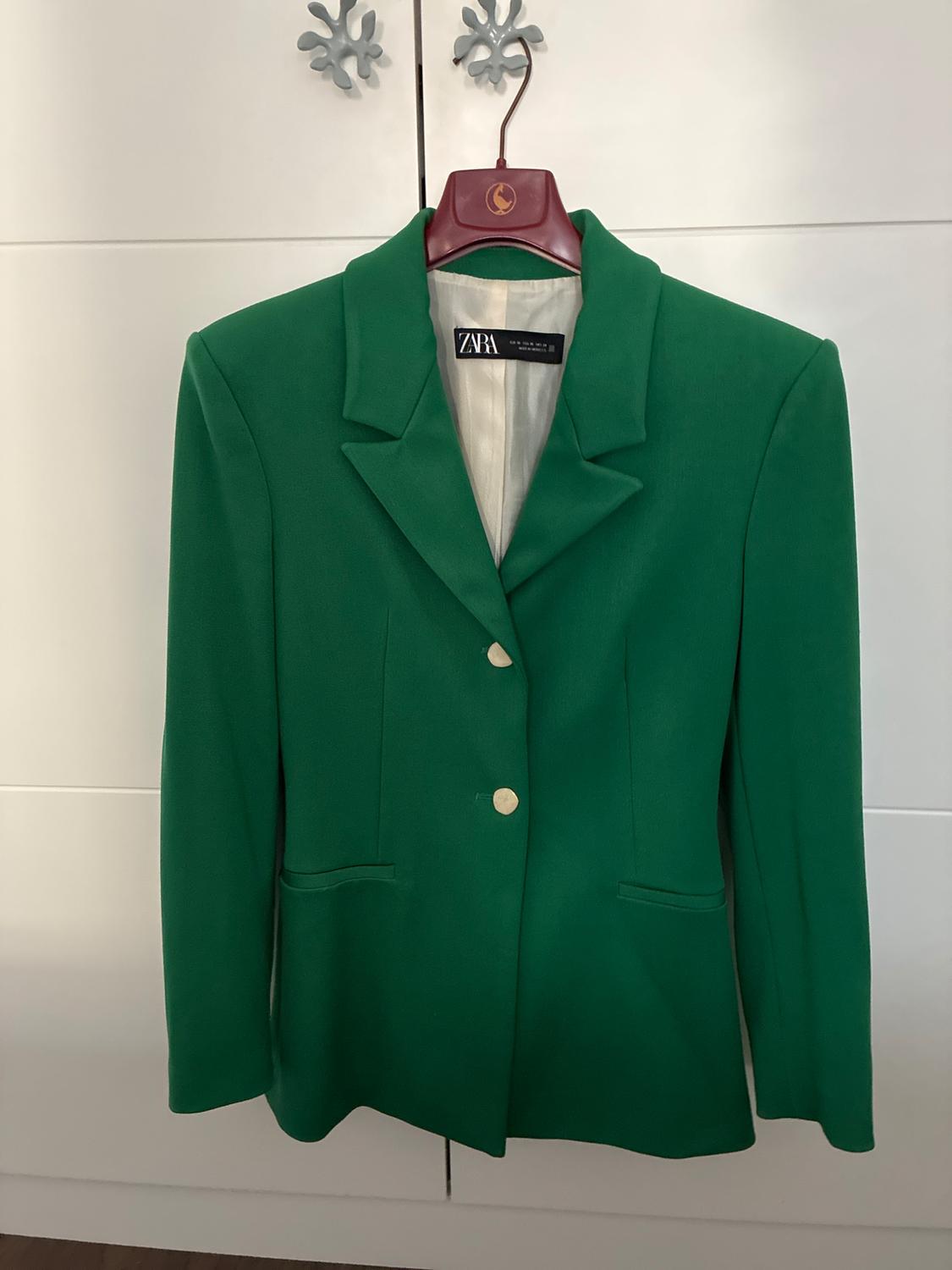 Green blazer with lapel collar and two buttons, hanging on a hanger against a white wardrobe. Interior label with the text ZARA.