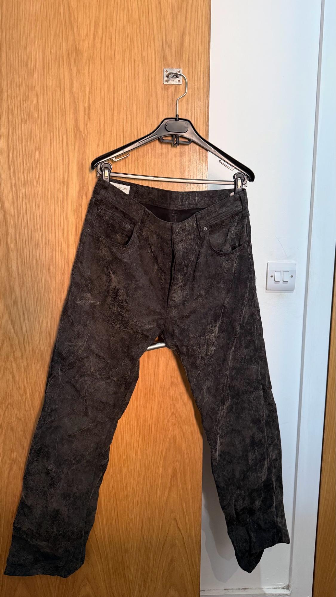 Textured dark trousers hanging on a hanger on a wooden door. Relaxed setting with a white wall and a light switch.