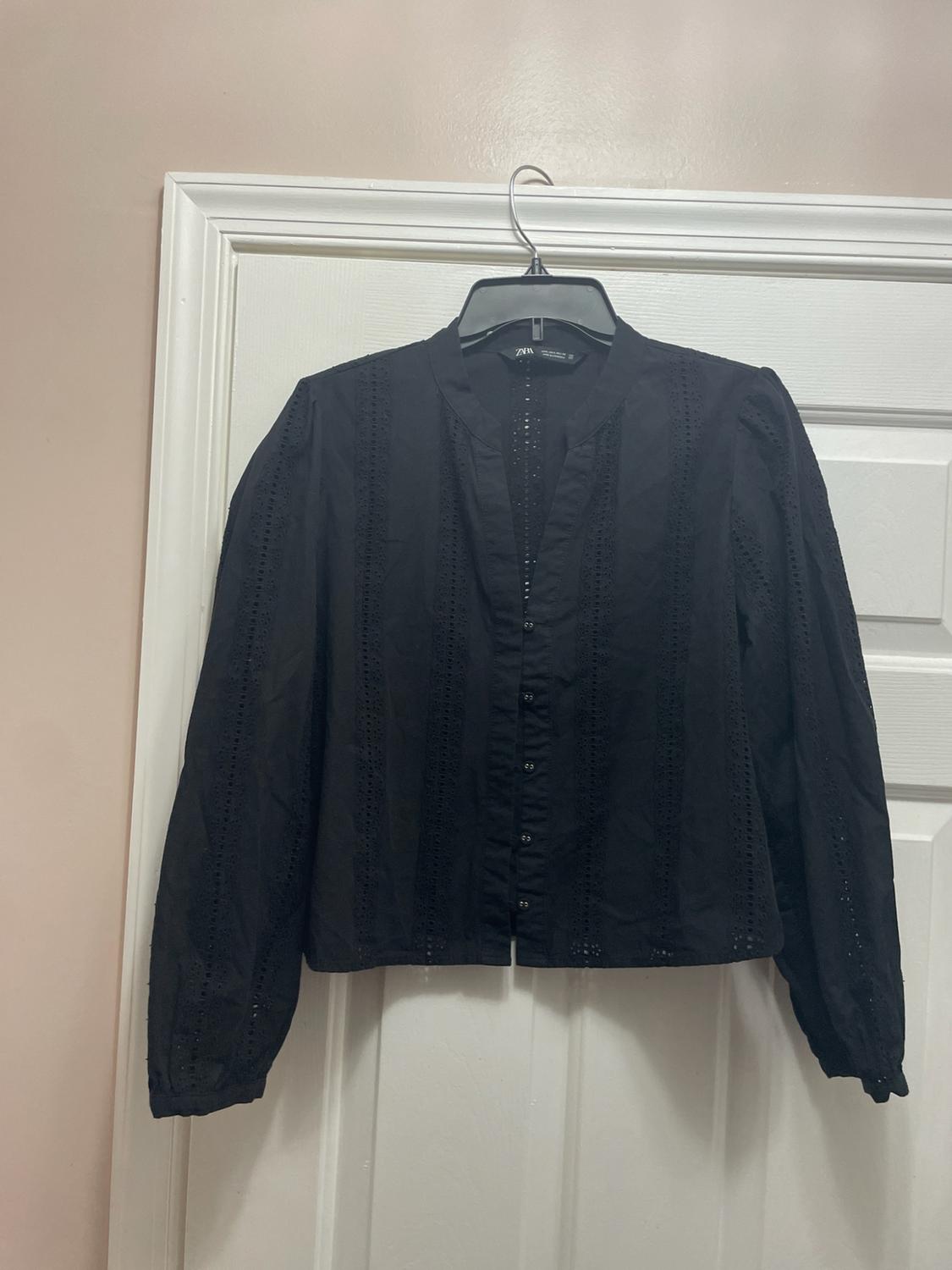 Black long-sleeve blouse with perforated details and front buttons, hanging on a white door. Label with the text ZARA visible.