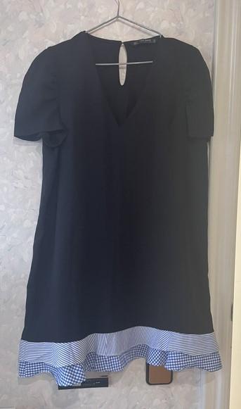 Black short-sleeved dress hanging on a hanger against a light wall. V-neck and ruffled hem with blue stripes and checks.