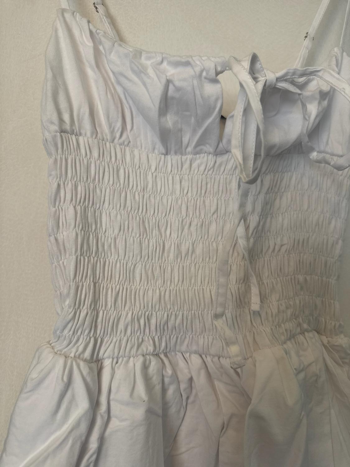 Close-up of a white garment with thin straps, a bow neckline, a stretch gathered bodice and a frilled hem against a light background.