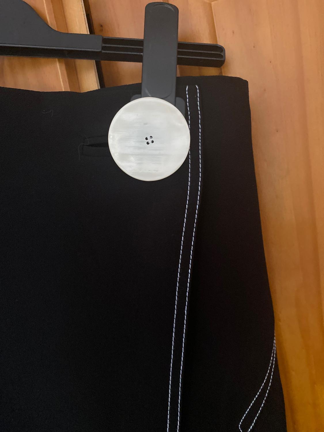 Black trousers with white stitching hanging on a black hanger against a wooden background. Large white button visible.