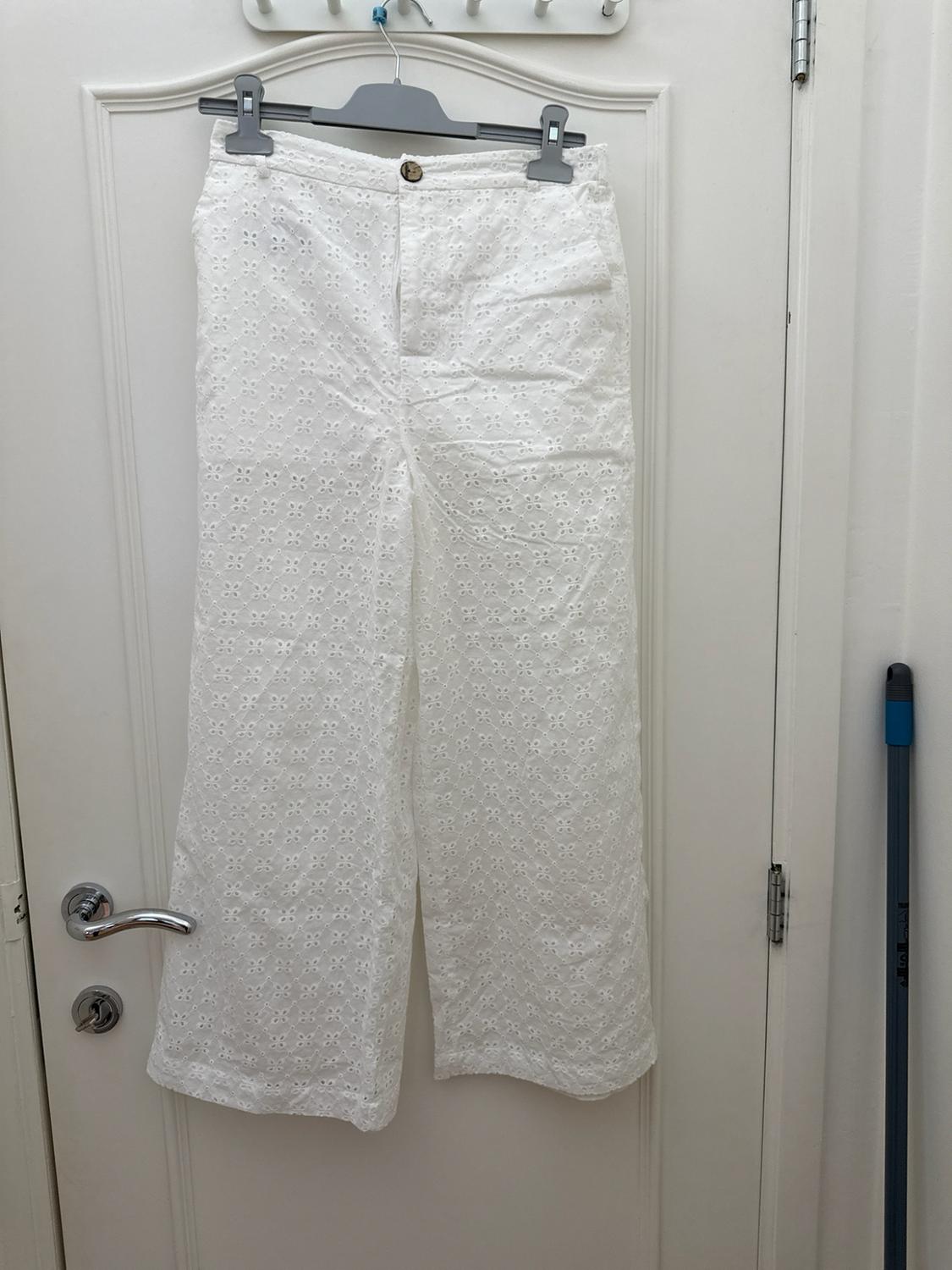 Wide white trousers with openwork embroidery, hanging from a clothes hanger on a white door with a silver handle.