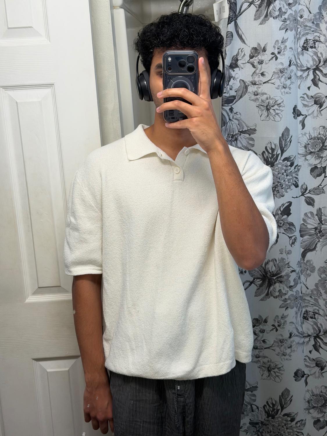 Man taking a selfie in a bathroom. He is wearing a white short-sleeved polo shirt, dark striped pants, and black headphones. Casual style.