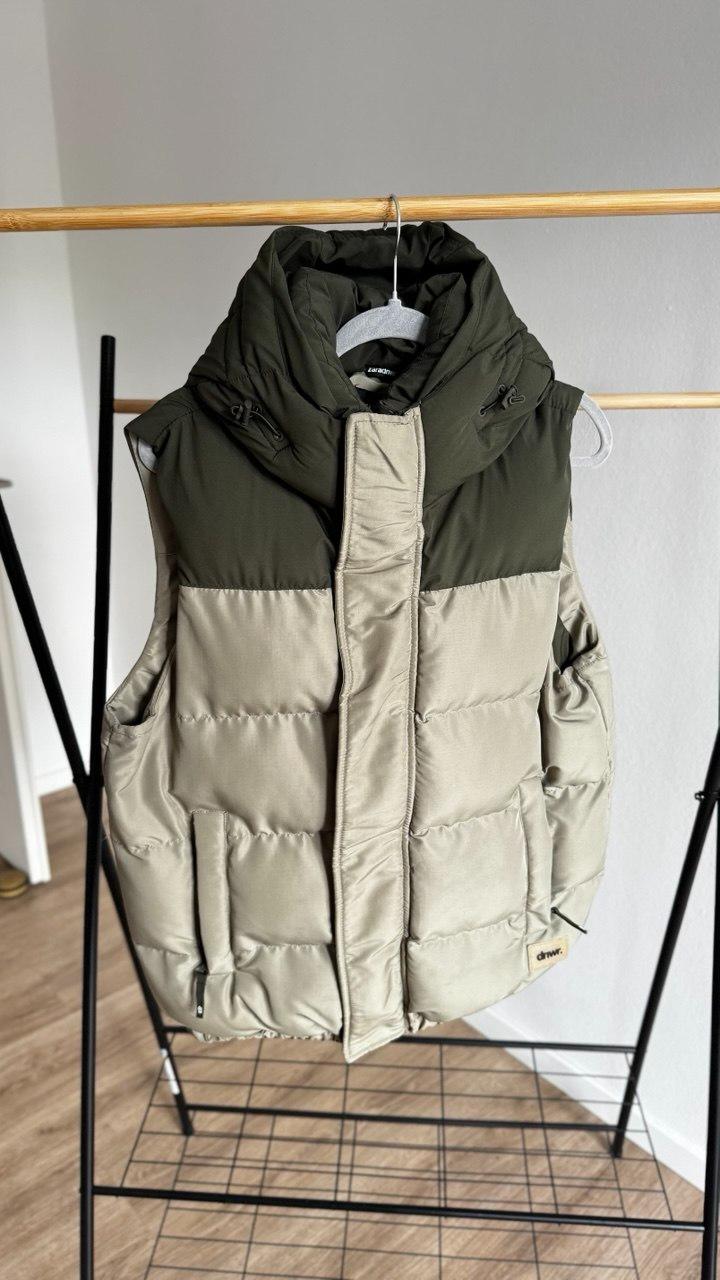 Dark green and beige quilted gilet on a wooden hanger and black coat rack. Relaxed style. Text: dnwr.