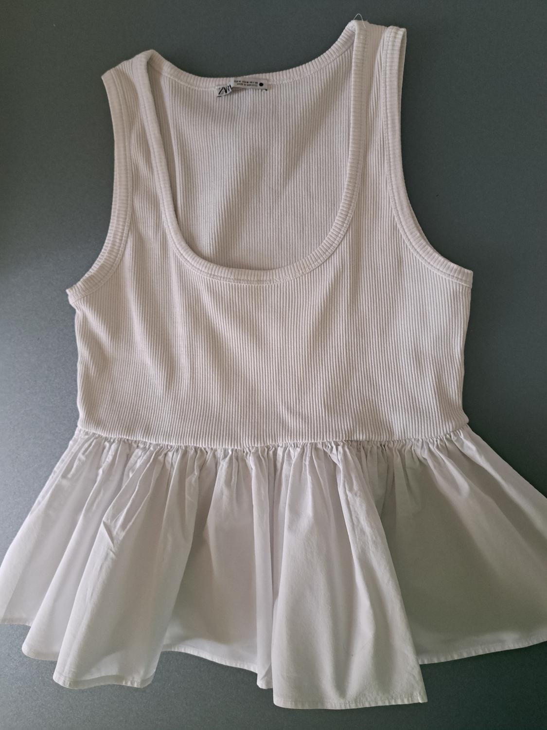 White sleeveless top with a ribbed knit upper and a ruched skirt lower, against a grey background.