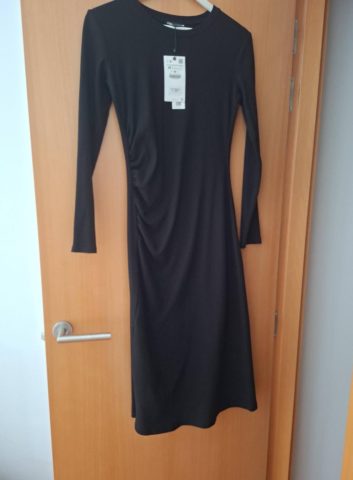 Black long-sleeved midi dress with a round neckline and ruched detail on one side. It is hanging on a light wooden door.