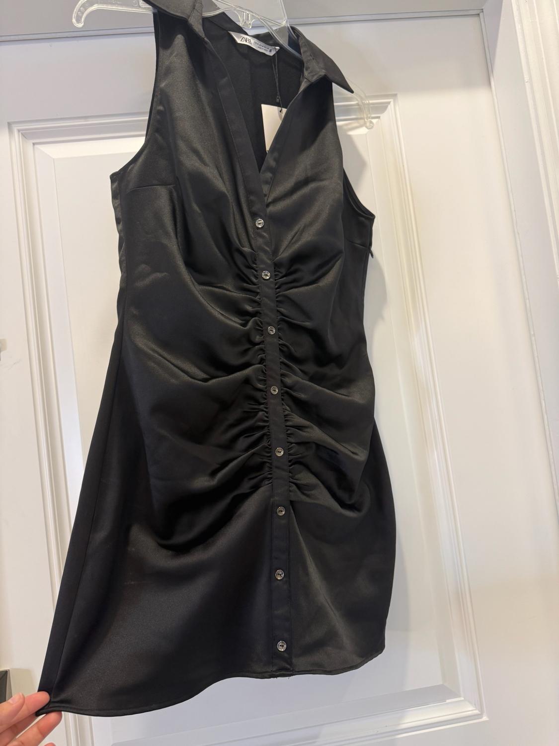 A sleeveless black dress featuring a collar, buttons and gathered detailing at the front is hanging over a white door. The ZARA label is visible, along with a hand holding the hem.