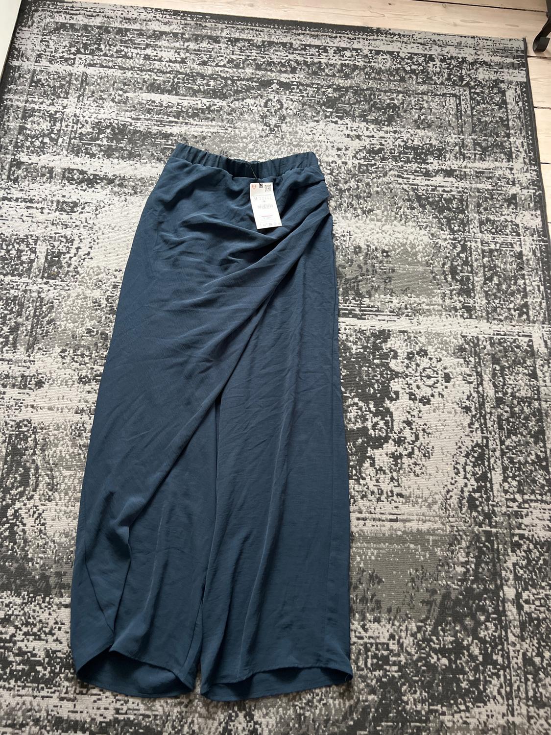 Loose blue trousers with a label, on a grey rug with an abstract design on a light wooden floor.