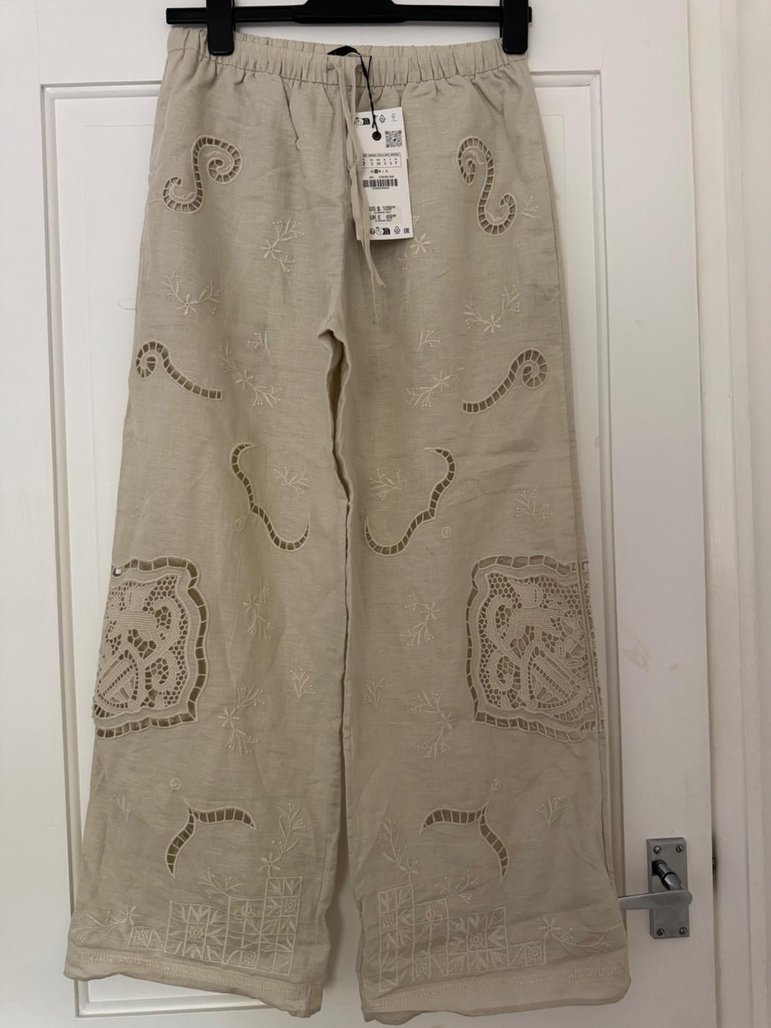 Beige wide-leg trousers with an elasticated waist and broderie anglaise details, hanging on a white door. Text: US $ 109 UK £ 69.
