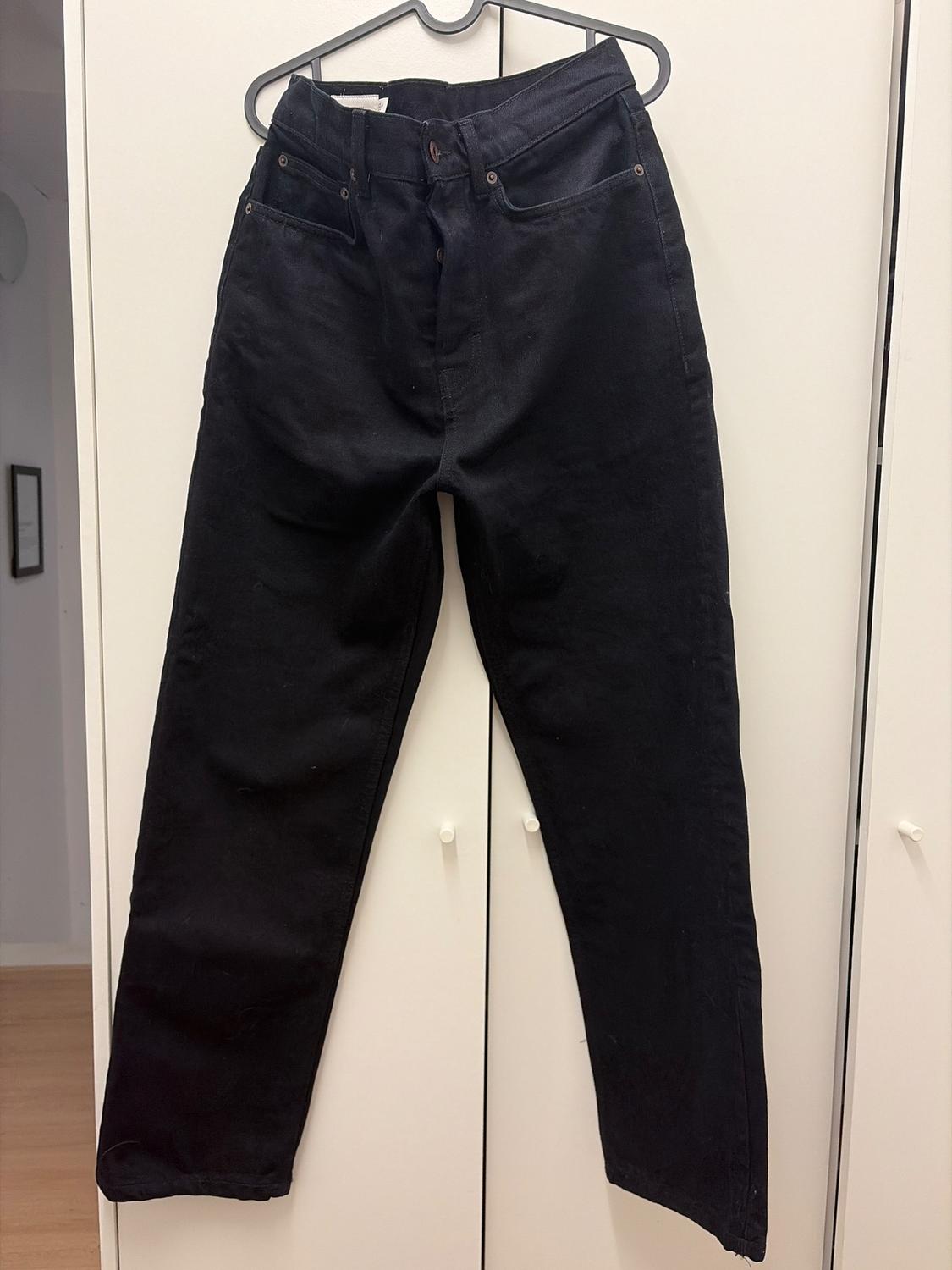 Black trousers hanging on a hanger in front of a white wardrobe. The room has light wooden flooring.