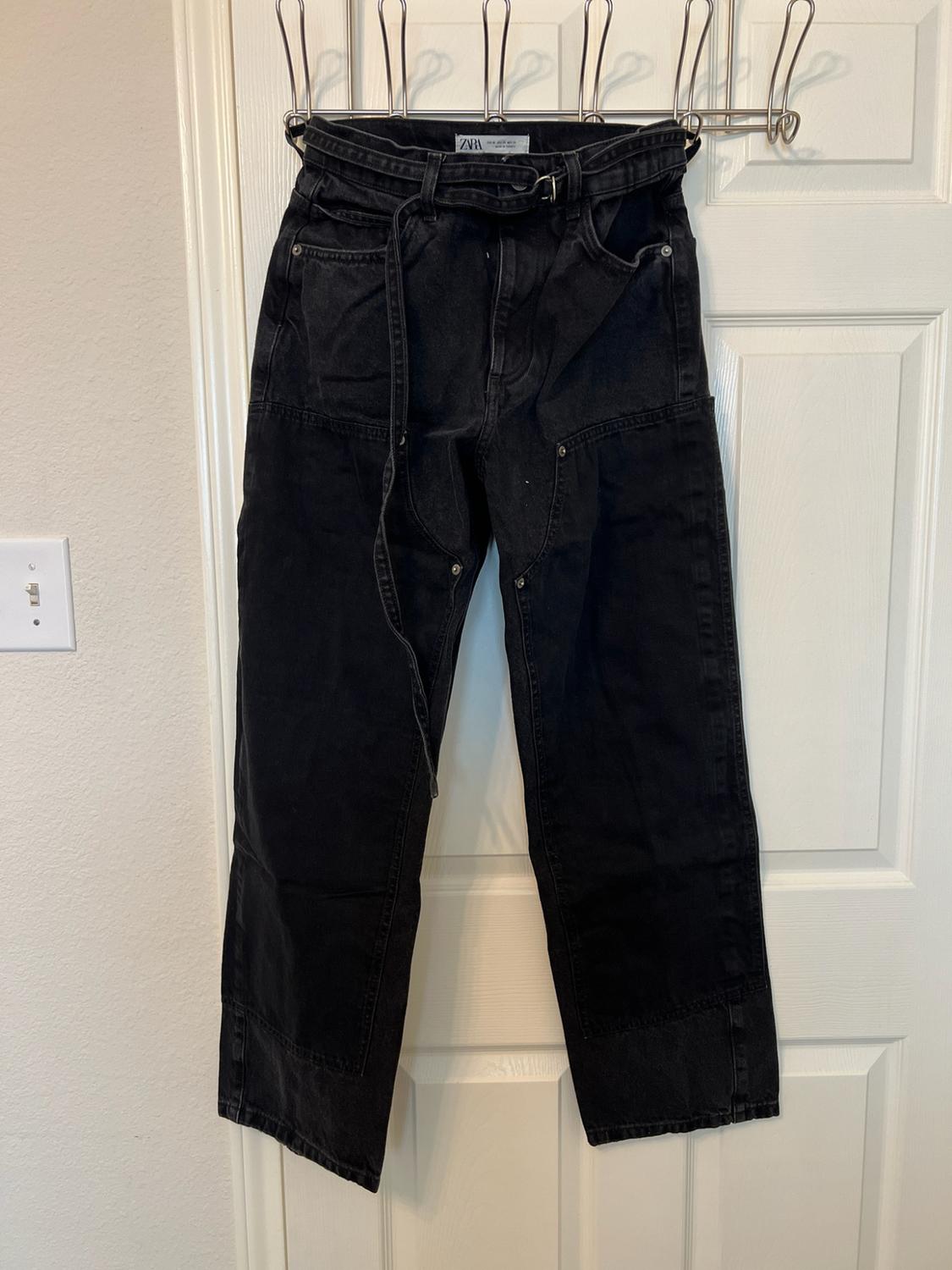 Black denim trousers with panels and a belt hanging on a white door. Label bearing the text ZARA and metallic details.