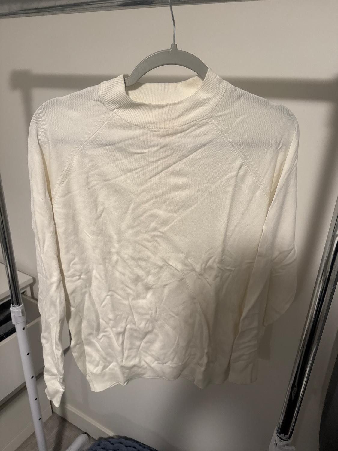Creased white jumper hanging on a grey hanger, against a neutral background. The garment has a crew neck and long sleeves.