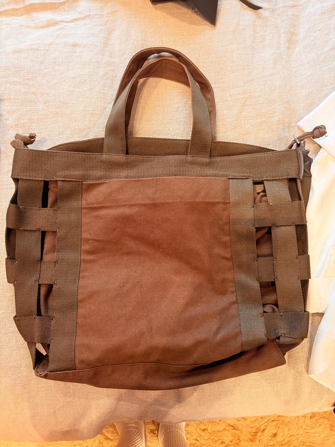 Brown fabric bag with short handles on a light surface. Design with interwoven straps on the sides.