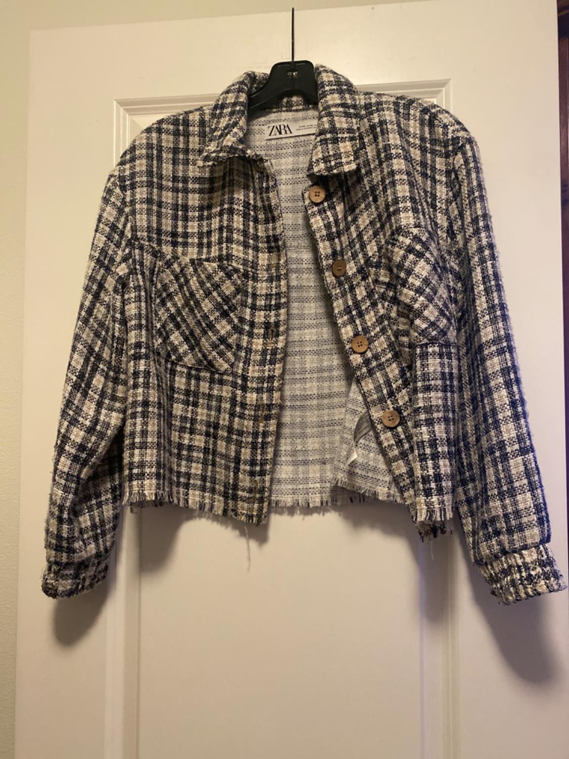Black and white checked cropped jacket with brown buttons, hanging on a black hanger over a white door.