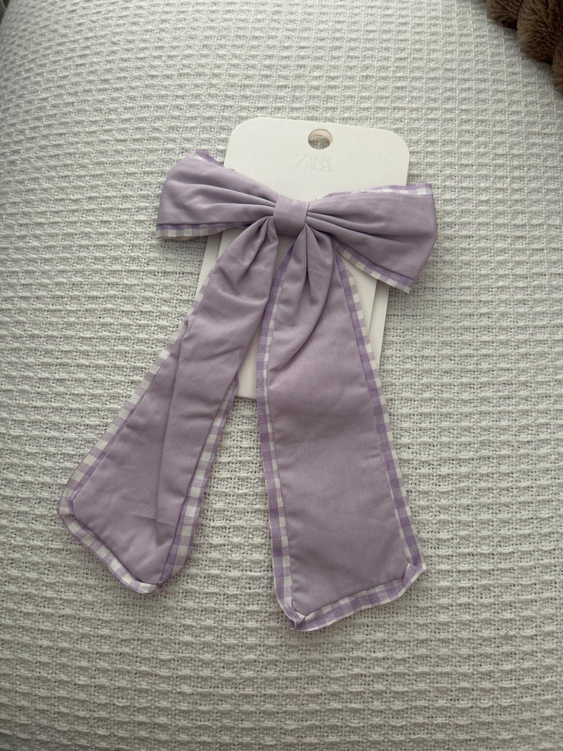 Lilac bow with checkered edges on a textured white background. Label with "ZARA" text barely visible.