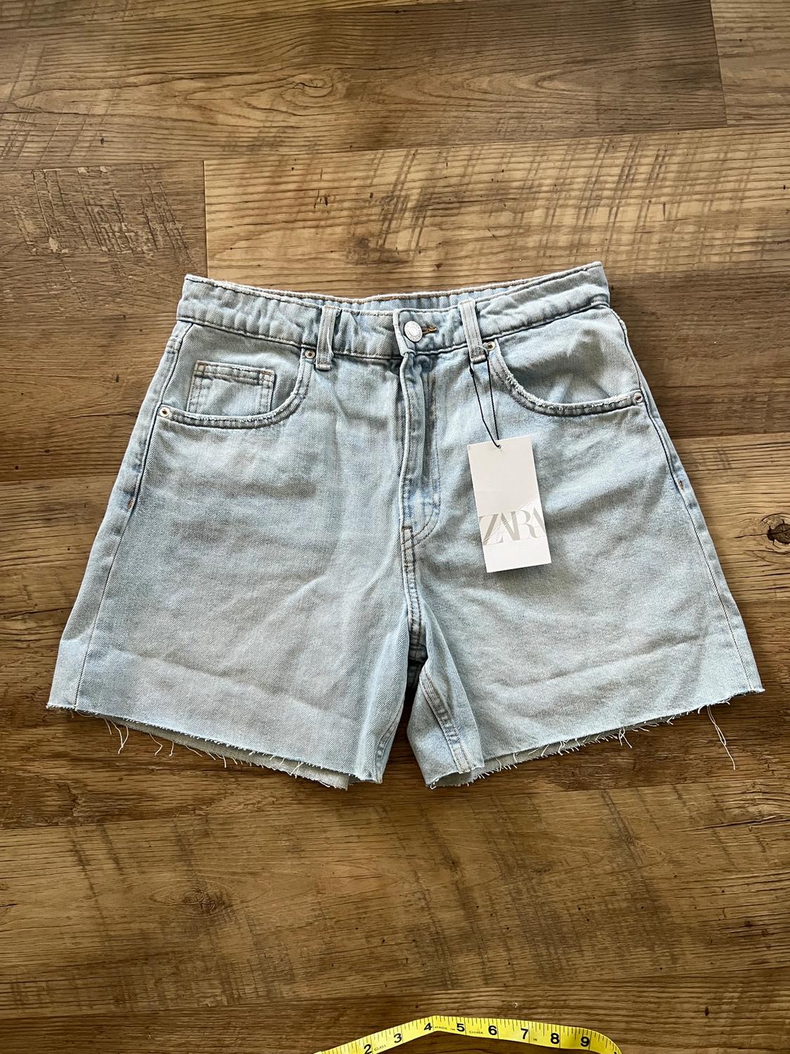 Light blue denim shorts with frayed hem on a wooden floor. Visible "ZARA" tag. Yellow tape measure at the bottom.