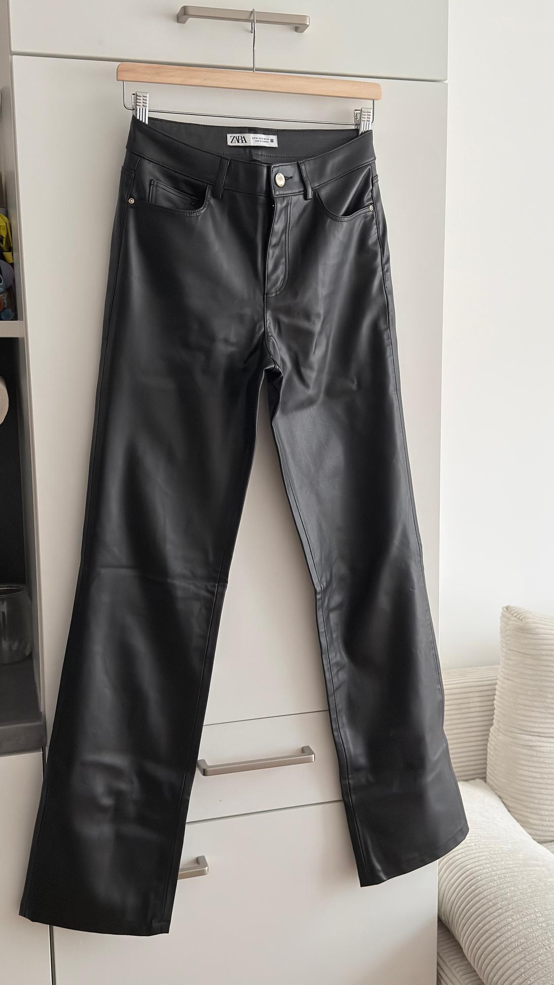 Black trousers hanging on a wooden hanger in front of a white wardrobe. Visible label with the text "ZARA".