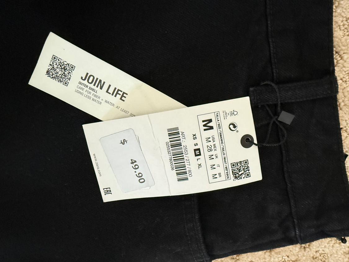 Black trousers label with text "JOIN LIFE", size M, price $49.90. Dark fabric background and beige carpet. Visible QR code.