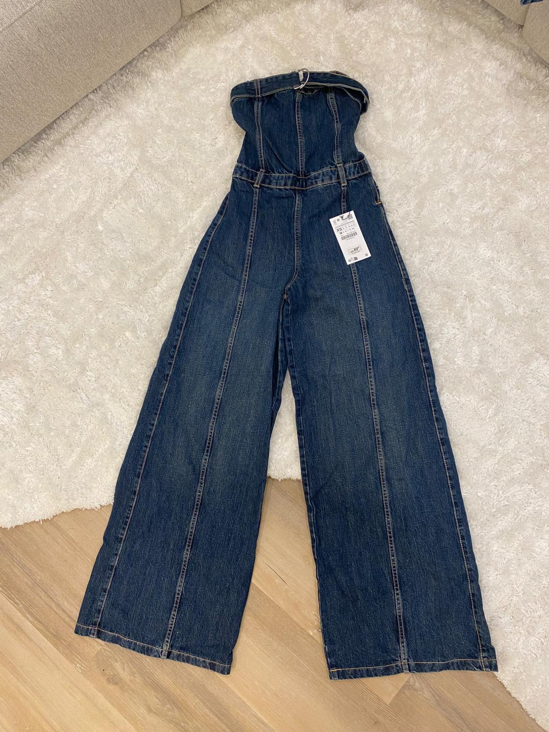Wide-leg denim jumpsuit with tag, on a white rug and light wood floor, next to a beige sofa.
