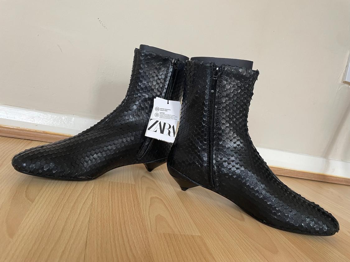 Black boots with a scale-like texture and a side zip, on a light wooden floor. Visible label with the text "ZARA".