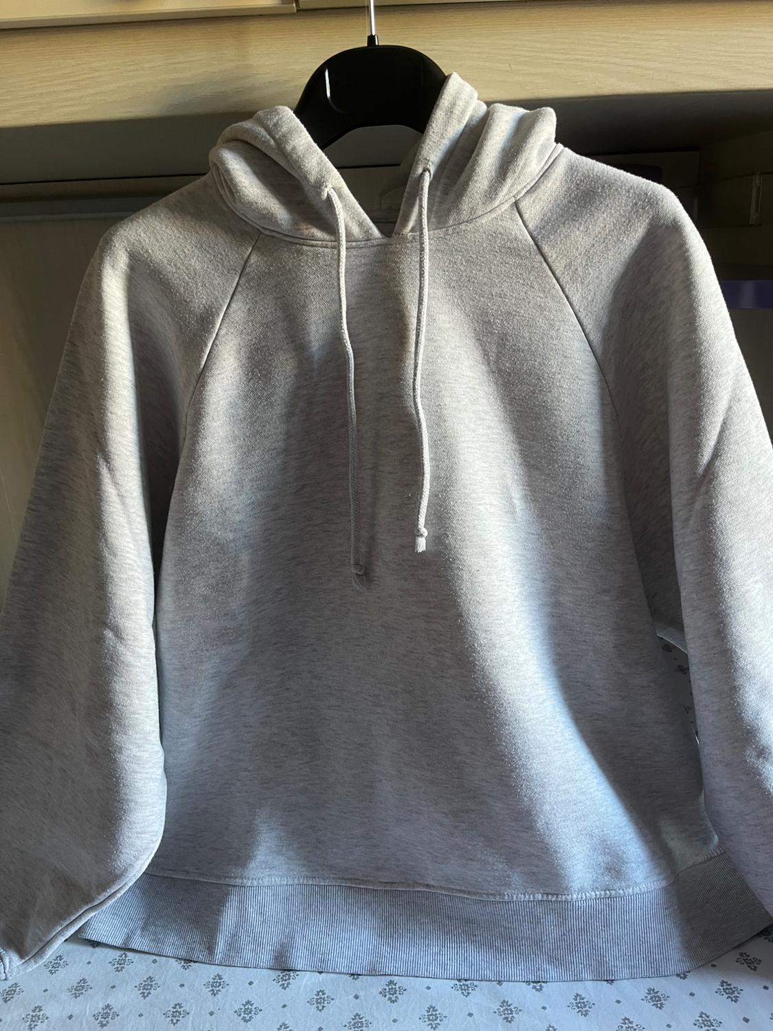 Light grey hoodie with drawstrings and raglan sleeves, hanging on a black hanger. Natural light, background with wooden shelf and patterned fabric.