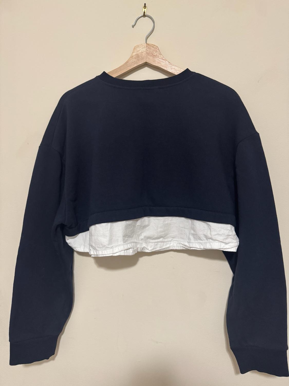 Cropped navy blue sweatshirt with a contrasting white hem, hanging on a wooden hanger against a light background. Casual and relaxed style.