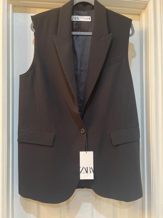 Black sleeveless vest hanging on a hanger, with lapels and one button. Visible tag with the text "ZARA". White door background.