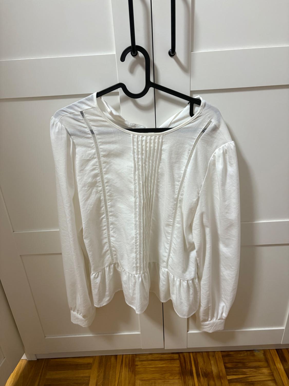 Long-sleeved white blouse with pleats and frills, hanging on a black hanger in front of a white wardrobe and wooden floor.