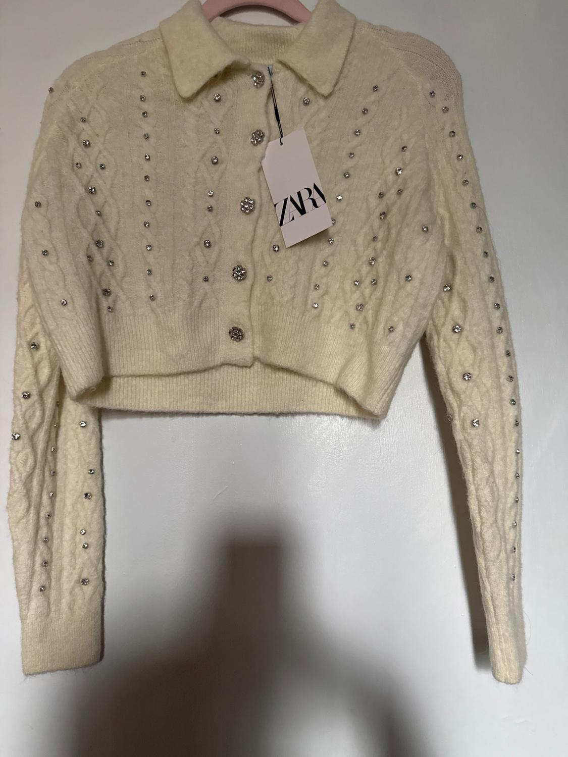 Cream knit cropped jacket with buttons and sparkly embellishments, hanging on a pink hanger. Label with text "ZARA".