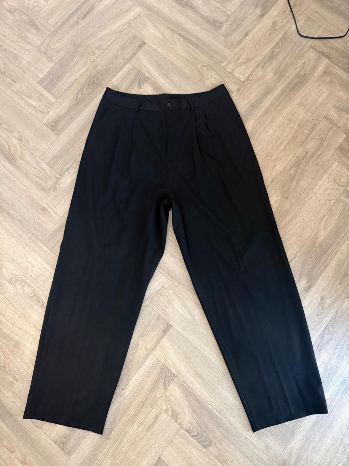 Black wide-leg trousers with pleats and a button, laid out on a herringbone wooden floor.
