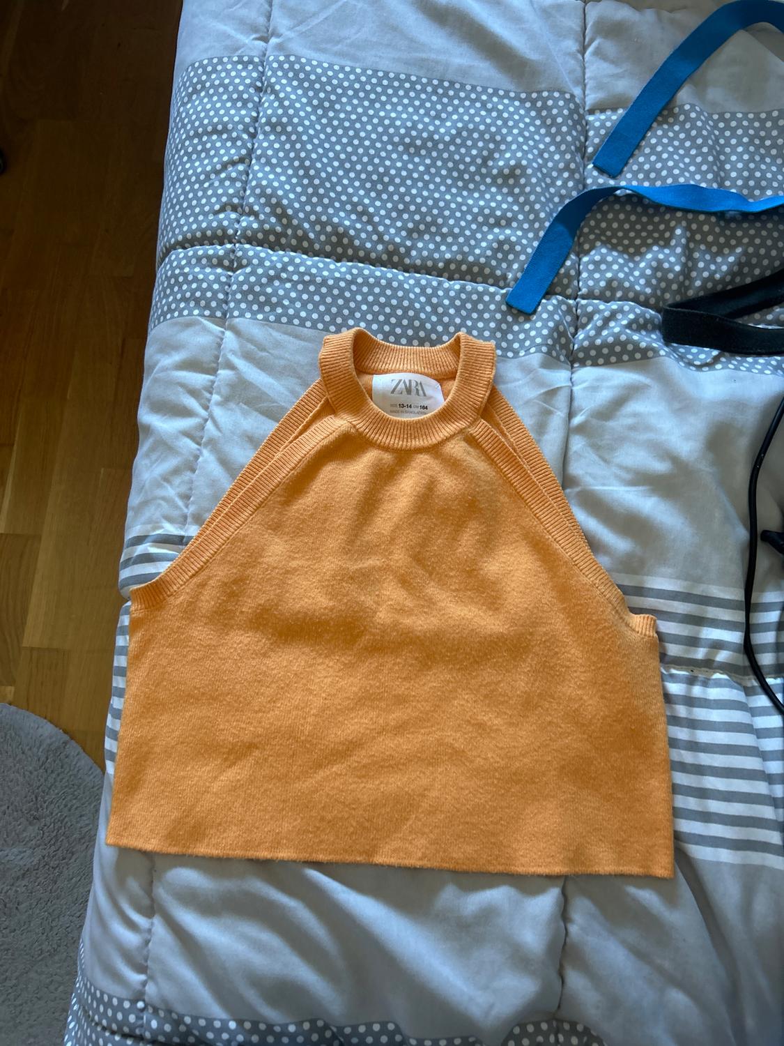 Orange sleeveless top on a grey quilt with white polka dots. Visible label with brand and size. Blue ribbons next to it.