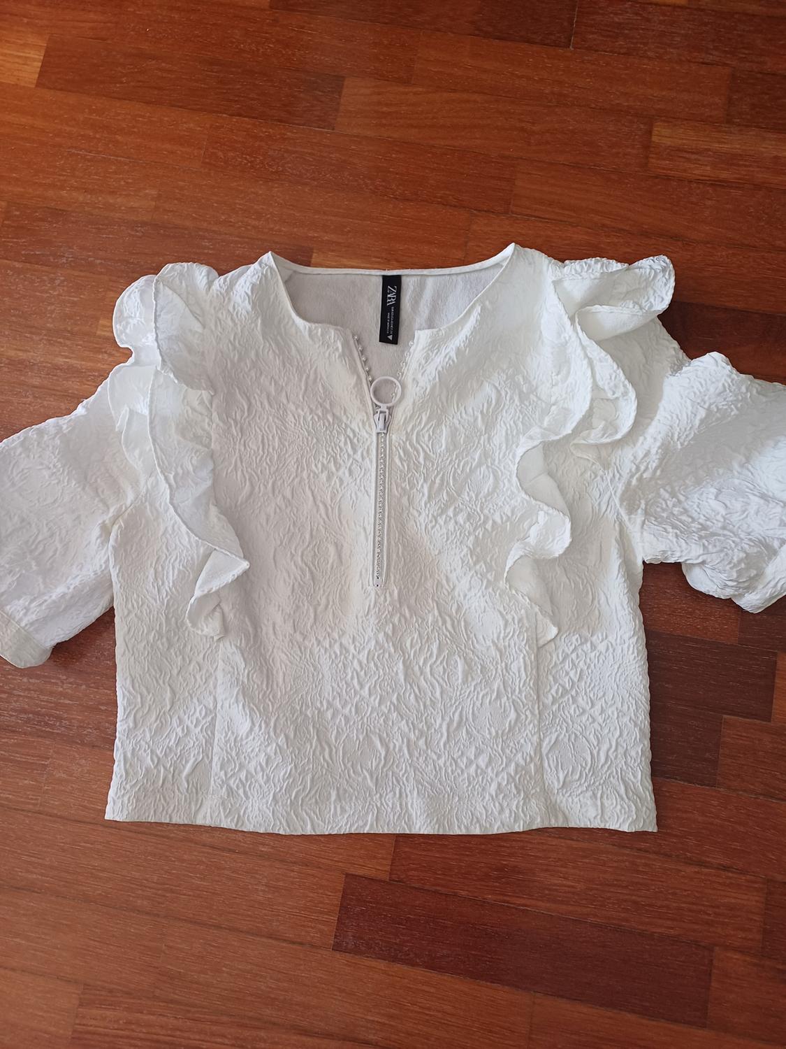Front view of a textured white blouse with a central zip and ruffled sleeves.